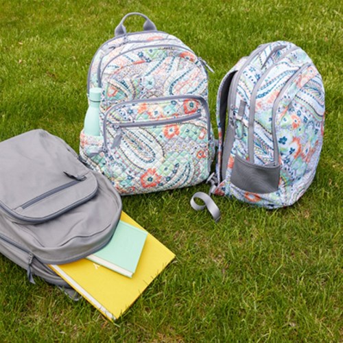 Two colorful backpacks and a gray bag with notebooks and a water bottle on green grass.