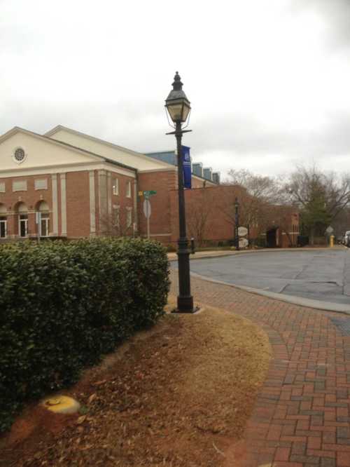 A street lamp beside a brick pathway, with a building and hedges in the background on a cloudy day.