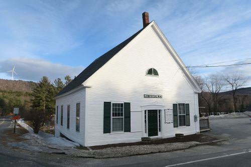 A white, gabled building with green shutters, located in a rural area with mountains in the background and a wind turbine.