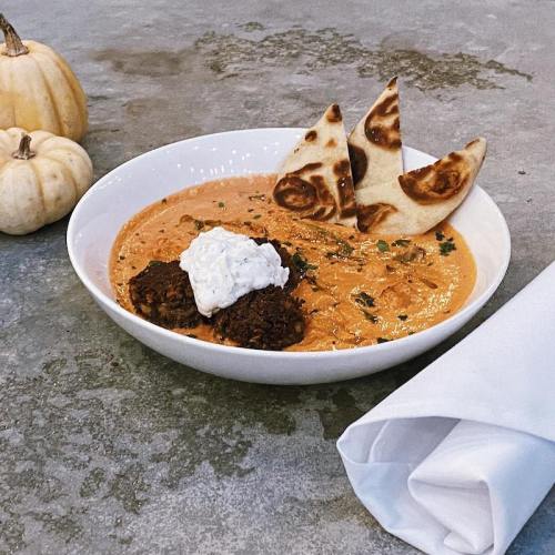 A bowl of creamy curry with meatballs, topped with yogurt and served with naan, alongside small pumpkins.