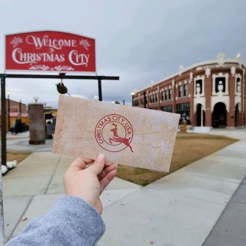 A hand holds a postcard in front of a "Welcome to Christmas City" sign and a historic building.