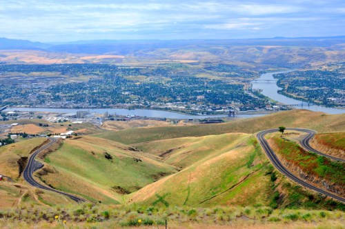 A panoramic view of rolling hills and a river winding through a valley, with a city in the distance under a cloudy sky.