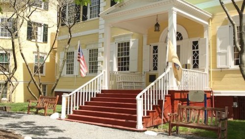 A yellow building with white trim, featuring a front porch, steps, and American flags, surrounded by benches and trees.