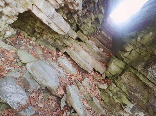 A rocky passageway with uneven stone steps, surrounded by moss-covered walls and scattered leaves. Light shines from above.