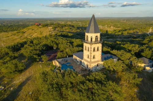 Aerial view of a tall, stone tower surrounded by greenery and a pool, set against a hilly landscape under a blue sky.