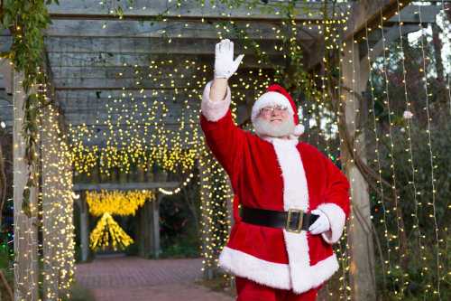 Santa Claus waves cheerfully in a festive, illuminated garden adorned with twinkling lights.