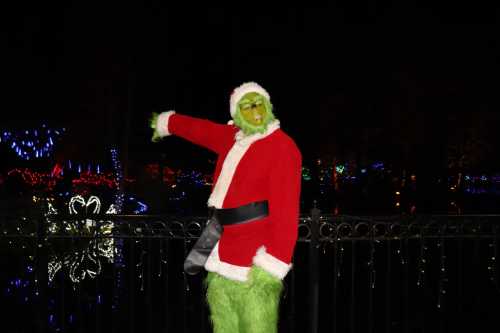 The Grinch in a Santa suit points playfully, surrounded by colorful holiday lights at night.