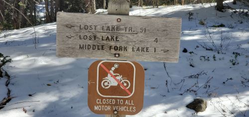 Wooden signpost in snowy forest indicating directions to Lost Lake and Middle Fork Lake, with a no motor vehicles symbol.