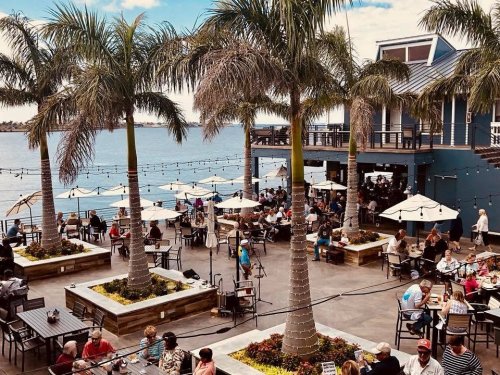A lively outdoor dining area by the water, featuring palm trees, umbrellas, and many people enjoying their meals.