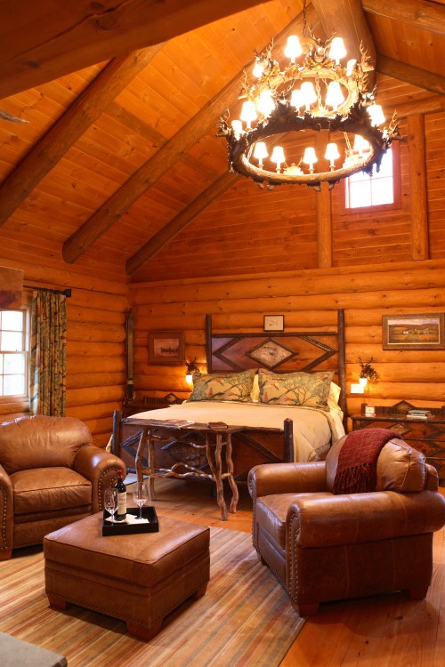 Cozy log cabin interior featuring a wooden bed, leather chairs, a chandelier, and warm, rustic decor.