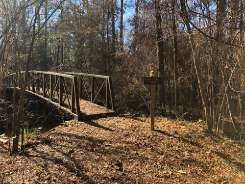 A wooden bridge crosses a small creek in a wooded area, with a sign nearby indicating a trail.