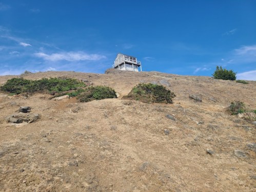 A small lookout tower sits atop a rocky hill under a clear blue sky, surrounded by sparse vegetation.