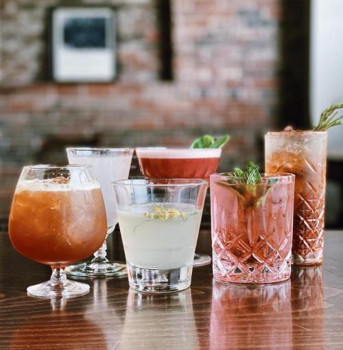 A variety of colorful cocktails in elegant glasses arranged on a wooden table against a brick wall backdrop.