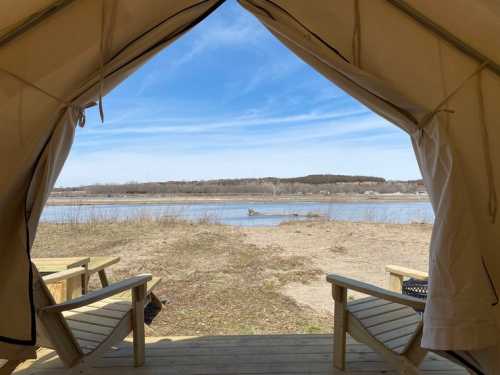 View from a tent opening, showcasing a serene river landscape with two chairs on a wooden deck.