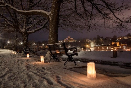 A snowy park at night with glowing lanterns lining a path, benches, and a view of a lit-up city in the background.