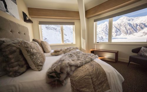 Cozy bedroom with a plush bed, fur throws, and large windows showcasing snowy mountain views.