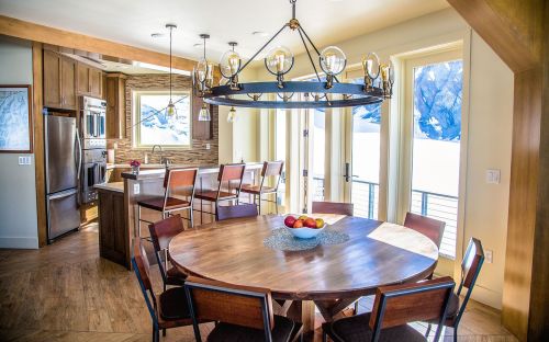 A modern kitchen and dining area with a round wooden table, chairs, and large windows overlooking a scenic view.