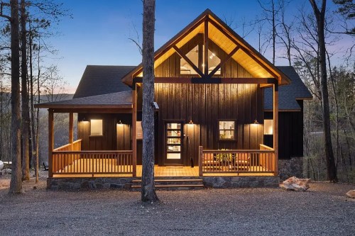 A modern wooden cabin surrounded by trees, featuring a porch and large windows, illuminated at dusk.