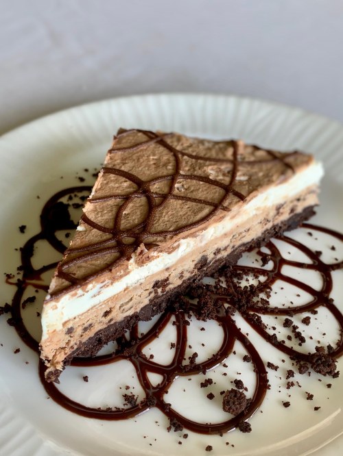 A slice of layered chocolate mousse cake on a plate, drizzled with chocolate sauce and surrounded by cookie crumbs.