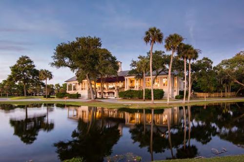 A beautiful house with large windows reflecting in a calm pond, surrounded by palm trees at dusk.