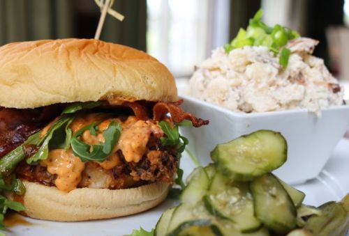 A close-up of a crispy chicken sandwich with bacon, lettuce, and sauce, served with potato salad and pickles.