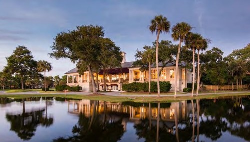 A charming house with large windows, surrounded by palm trees, reflecting in a calm pond at dusk.