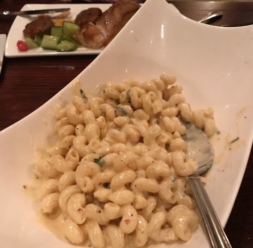 A bowl of creamy macaroni pasta with a spoon, served on a wooden table with a plate of meat and vegetables in the background.