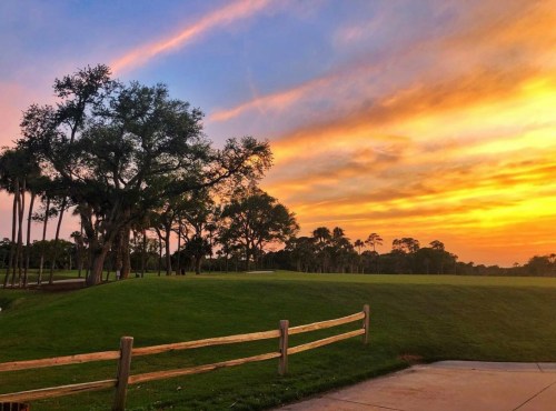 A vibrant sunset over a golf course, with trees and a wooden fence in the foreground.