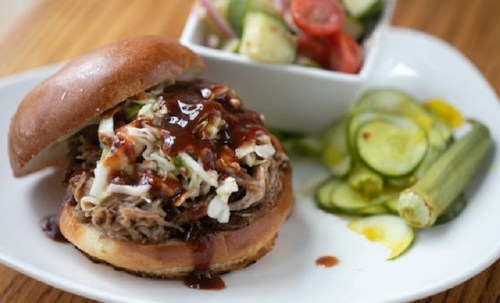 A pulled pork sandwich topped with coleslaw and barbecue sauce, served with pickles and a side salad.