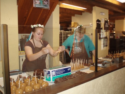 Two women in aprons prepare caramel apples at a counter, surrounded by various treats and a display of candy.