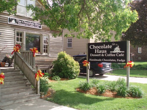 A cozy café named Chocolate Haus, featuring a sign and decorative bows, surrounded by greenery and a parked car.