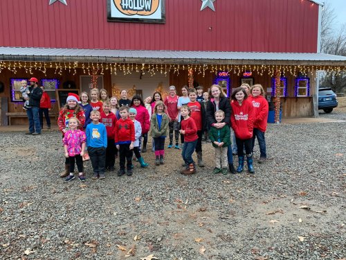 A group of children and adults in festive attire pose outside a decorated building with holiday lights.