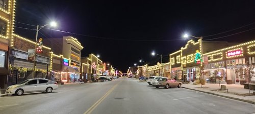 A brightly lit street at night, lined with festive lights and decorated storefronts. Cars are parked along the road.
