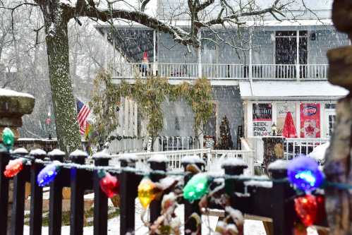 A snowy scene featuring a house decorated with colorful lights and an American flag, surrounded by falling snow.