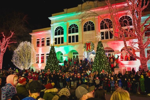 A festive crowd gathers in front of a beautifully lit building adorned with Christmas decorations and trees.
