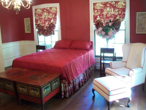A cozy bedroom with red walls, a red bedspread, and elegant furniture, including a white armchair and decorative curtains.