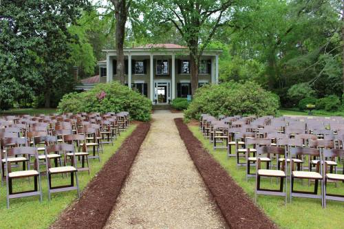 A scenic outdoor venue with rows of wooden chairs lining a gravel path leading to a large house surrounded by greenery.