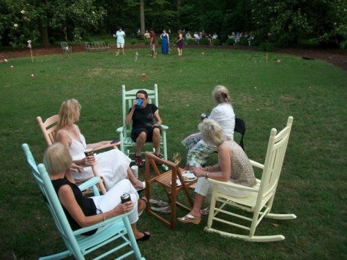 A group of women in rocking chairs enjoying drinks and conversation in a grassy outdoor setting.