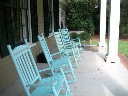 A porch with four light blue rocking chairs lined up in front of a house, surrounded by greenery.