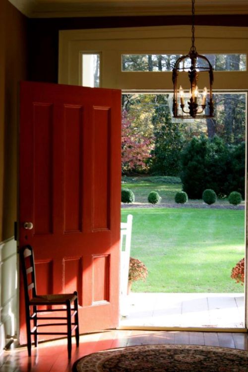 A red door stands open, revealing a lush green garden and a small chair inside the warmly lit entryway.