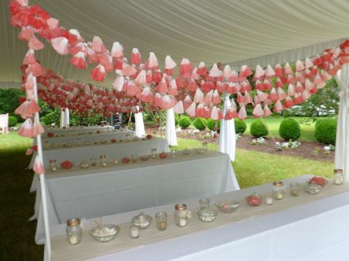 A tented outdoor space decorated with pink and white tassels, featuring tables set with candles and decorative items.