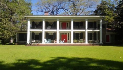 A large two-story house with white columns, red doors, and a green lawn surrounded by trees.