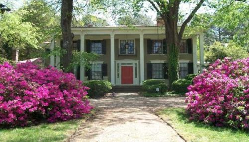 A charming two-story house with a red door, surrounded by vibrant pink azaleas and lush greenery.