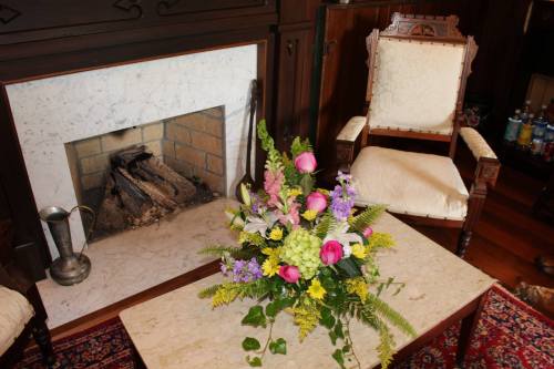 A cozy room with a floral arrangement on a table, a fireplace, and an ornate chair in the background.