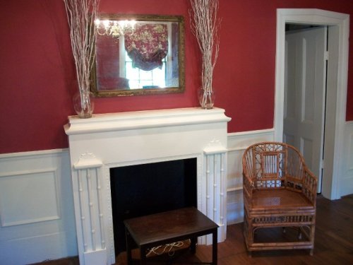 A cozy room with a red wall, white fireplace, mirror, and a wicker chair beside a small table.