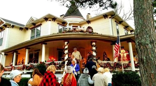 A festive house decorated for Christmas, with people gathering on the porch and holiday lights adorning the exterior.