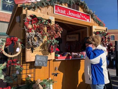 A wooden market stall decorated with wreaths and holiday items, with a customer browsing in a blue jacket.