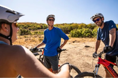 Three mountain bikers in helmets chat and smile in a sunny outdoor setting with greenery in the background.