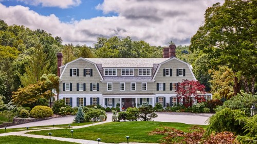 A large, elegant house with green shutters, surrounded by lush gardens and trees under a partly cloudy sky.