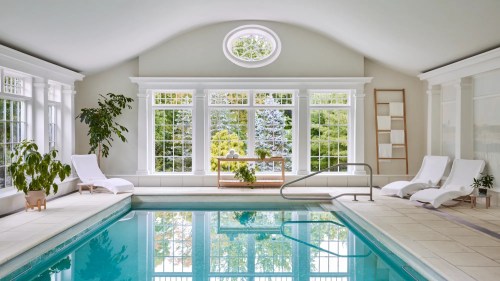 Indoor pool area with large windows, natural light, and lounge chairs, surrounded by greenery.